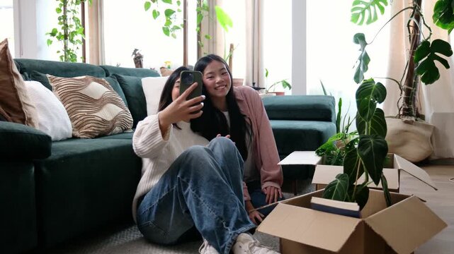 Two cheerful female roommates taking a selfie on a smartphone while sitting on the floor after moving in
