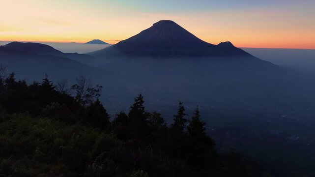 Aerial footage of Mount Sindoro set against a sunrise sky featuring golden and blue hues. The drone shot captures the majestic silhouette of the mountain before dawn on a clear morning.