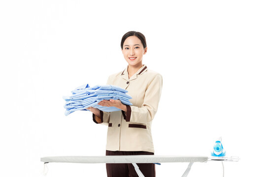 Smiling housekeeper holding folded laundry beside ironing board