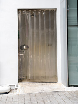 Emergency shower and eye wash station behind transparent plastic strip curtains in a white wall alcove. Safety equipment for industrial or lab environments to prevent chemical injuries.