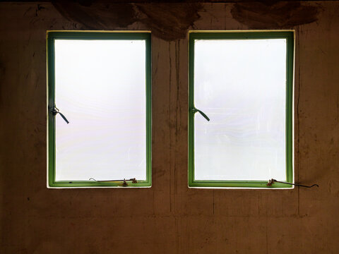 Two green frame windows on a textured grunge concrete wall. Bright sunlight through glass. Interior of an old or unfinished industrial building. Minimalist composition with architectural detail.