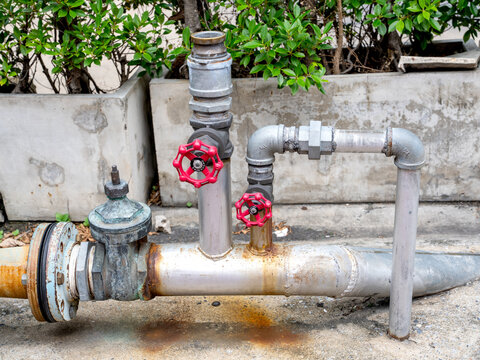 Large galvanized steel water pipe system with red shut-off valves and elbow joints. Outdoor industrial plumbing infrastructure near concrete planters and greenery. Professional utility equipment.