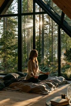 young woman meditating by large window overlooking forest, peaceful mindfulness meditation in scandinavian minimalist home, serene morning meditation with forest view in modern interior