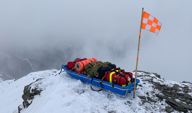  Bright blue mountain rescue toboggan loaded with emergency gear parked near a ski patrol marker flag on a foggy cliff edge, High-visibility colors against soft grey fog, wind-dusted