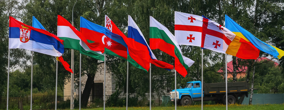 Multiple flags flying in a row in a field during a clear day with green trees in the background