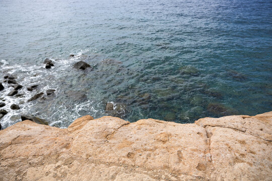 Der Blick von einer felsigen Klippe hinunter auf das tiefblaue, klare Meer mit sanften Wellen, die an den Steinen im Wasser brechen. Eine idyllische K&uuml;stenlandschaft voller Ruhe und nat&uuml;rlicher Sch&ouml;nh