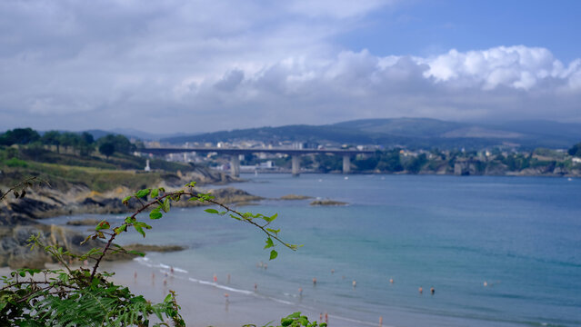 Scenic view of Playa de Arnao in Castropol, Asturias, featuring a picturesque sandy beach along the calm blue estuary waters, framed by lush coastal greenery and a distant bridge under a cloudy sky