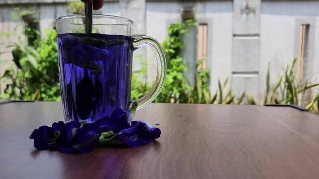 Person stirring a glass mug of deep blue butterfly pea flower tea with a spoon on a wooden table with fresh Clitoria ternatea flowers scattered around in an outdoor setting