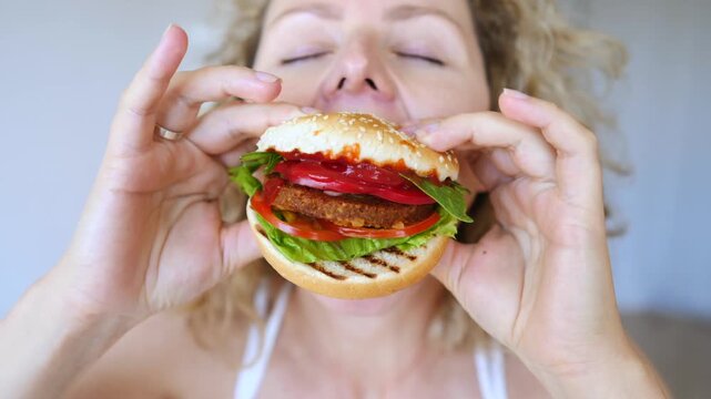 Close up of a happy blonde woman with curly hair bringing a delicious veggie burger to her mouth, enjoying the smell with her eyes closed, and preparing to take a big bite