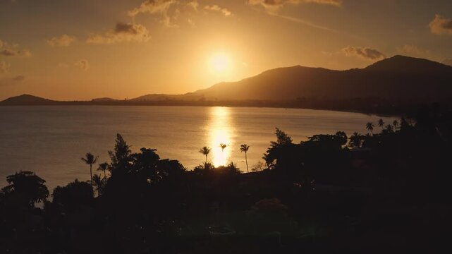Samui island tropical sunset landscape. Sun dipping below distant mountains, reflecting vibrant orange glow on serene water with silhouetted palm trees and lush tropical coastline. Aerial drone flight