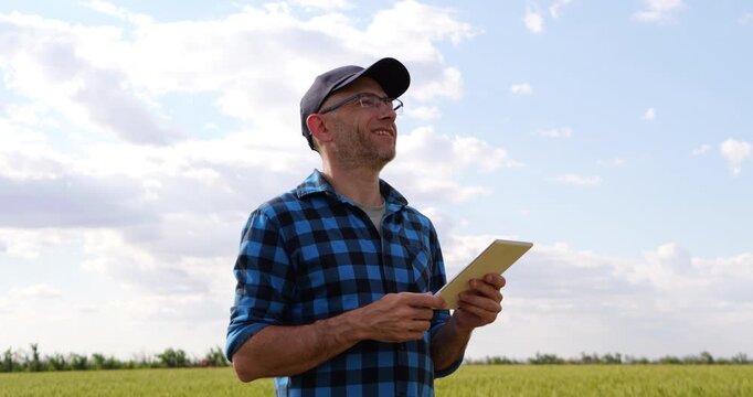 Man using tablet in green field under blue sky. Agronomist smiles while checking information on device. Bright atmosphere creates positive mood for outdoor technology use.