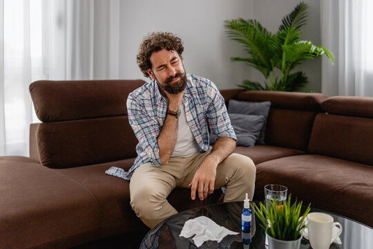 Man rubbing sore neck on couch at home feeling unwell with tissues and nasal spray nearby