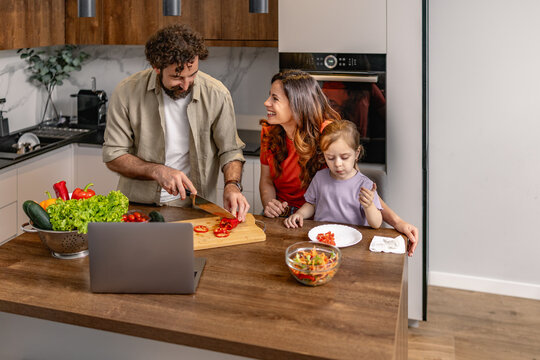 Family cooking together in modern kitchen with dad chopping vegetables while mom and daughter watch