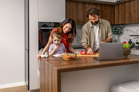 Family cooking together in modern kitchen with parents feeding child and following online recipe