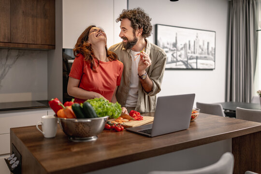 Couple cooking together in modern kitchen laughing while preparing healthy meal with laptop