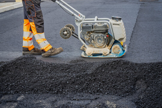 Construction worker operating plate compactor machine on fresh asphalt pavement surface during road paving and civil engineering infrastructure works