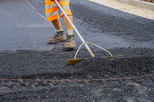 Construction worker using long handled asphalt lute to manually level hot mix asphalt surface layer during road paving and civil engineering works