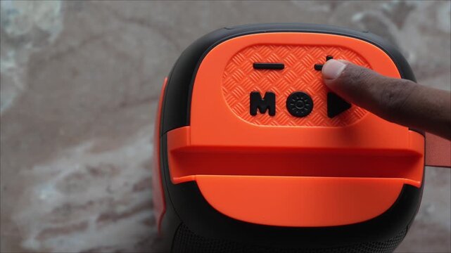 Hand increasing volume on an orange and black portable bluetooth speaker