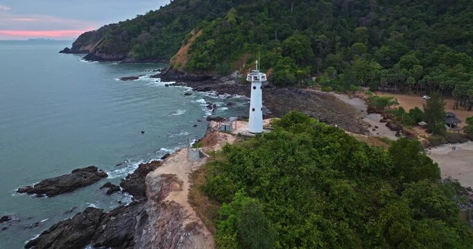 Lighthouse standing on a rocky cliff overlooking the ocean at Mu Ko Lanta National Park, Waterfall Bay, Thailand, dramatic sky at dusk providing navigation guidance for vessels. Aerial drone panorama
