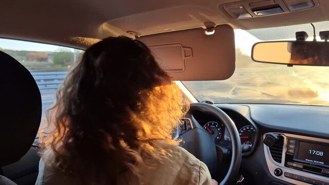Young woman driving car during sunset, adjusting sun visor while holding steering wheel on highway. Static interior shot from behind passenger seat.