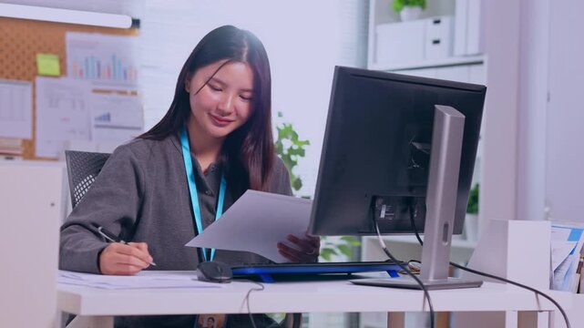 Asian businesswoman working at office desk with computer and document, professional woman writing notes, reviewing paperwork in modern workplace