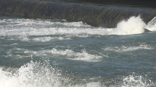 Dramatic slow motion shot showcasing smooth river water cascading off a rubber dam crest and crashing violently into dense white foam below.
