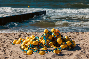 Pile of yellow fishing buoys on a sandy beach with rolling sea waves and a wooden breakwater in the background. © Dmytro