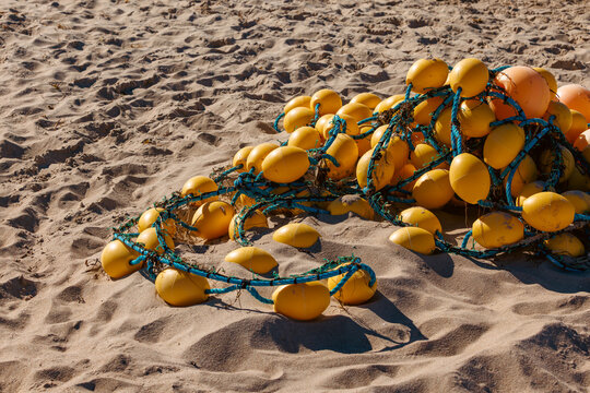 Tangled pile of bright yellow and orange fishing buoys linked by blue ropes on a textured sandy beach in the sun.