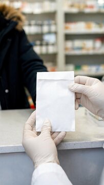 Pharmacist in white coat hands over a sealed white envelope to a customer at a pharmacy counter with shelves of medication in the background