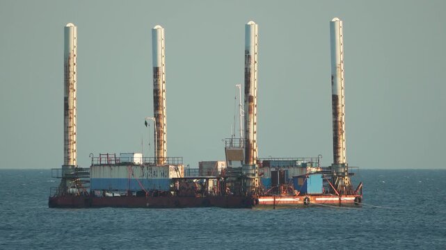 Offshore platform barge, rusty jack-up vessel floats on blue ocean under clear sky