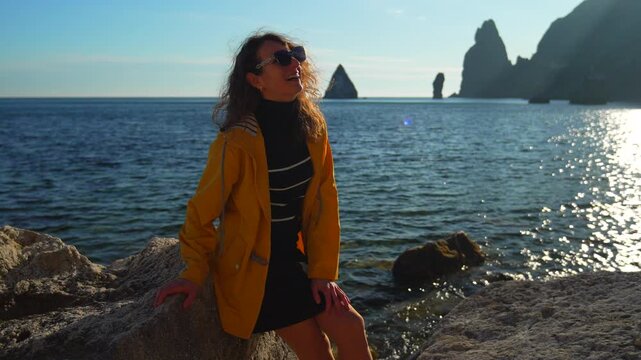 Traveler coastline sunshine young woman in yellow jacket sitting on coastal rocks looking at the ocean enjoying travel and freedom