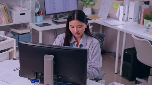 Asian woman office employee working at computer monitor on desk in modern workspace with document shelf chair plant device business technology professional focused mood