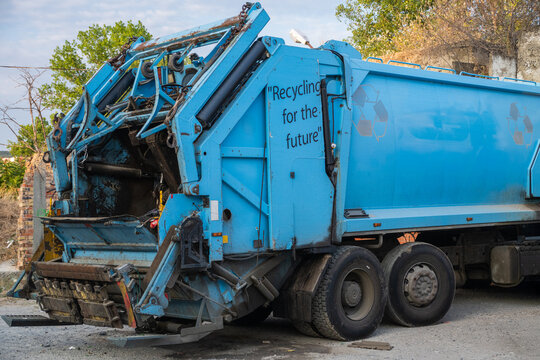 A heavy-duty, light blue garbage and recycling truck is parked outdoors with its complex rear hydraulic compactor mechanism exposed