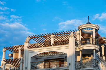 A luxurious white building exterior featuring a distinctive wooden pergola, classic columns, and a circular balcony with ornate iron railings against a partly cloudy blue sky © Andrey