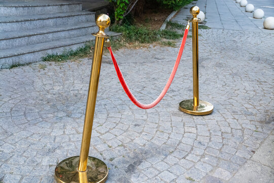 Two gold stanchions connected by a red velvet rope stand on an outdoor cobblestone surface, serving as a VIP entrance or crowd control barrier,