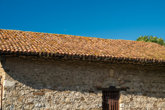An ancient stone building with a weathered terracotta tiled roof stands illuminated by bright sunlight under a clear, cloudless blue sky.