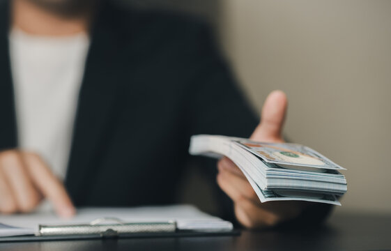 A professional person in formal attire holds a stack of currency bills and financial documents, indicating financial transactions or business activities in an office setting.
