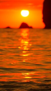 Burning orange sun sets behind rocky island silhouettes as gentle water surface ripples at dusk. Vertical static extreme close-up shot.