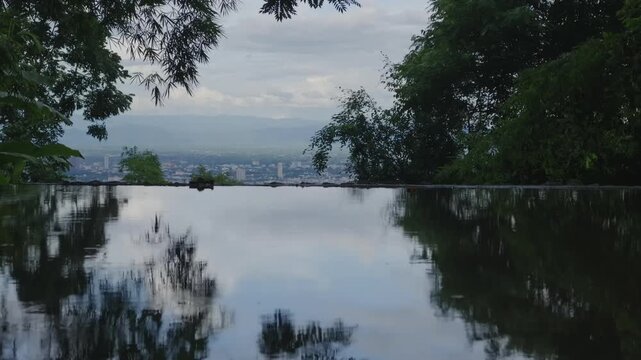 Panoramic city skyline reflected in calm water from forest viewpoint, Chiang Mai landscape with mountain horizon framed by tropical trees and cloudy sky 4k