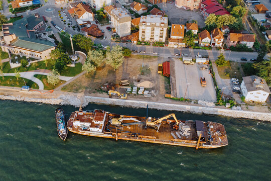 Rusty work barge moored along a riverside construction quay with cranes and machinery, overlooking a small European town from above.