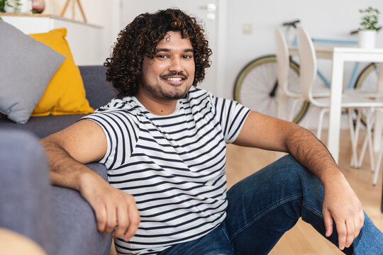 Young adult Indian man sitting on floor at home living room. Young Indian man smiling in comfortable apartment interior. Lifestyle and home concept.