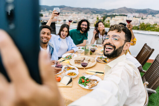 Diverse friends enjoying a celebratory party on an outdoor terrace, raising wine glasses while capturing a joyful selfie moment during a summer gathering