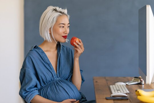 Pregnant woman eating a red apple at a home office desk for maternity wellness banner and healthy lifestyle background