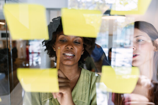 Diverse businesswomen collaborating on a project, organizing ideas on sticky notes placed on a glass wall, sharing insights and developing strategies in a modern office meeting