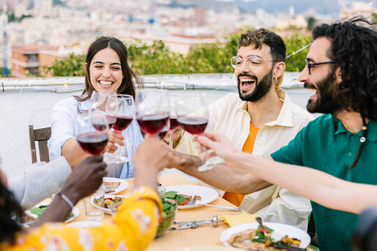 Diverse friends enjoying a celebratory barbecue gathering on a building terrace, clinking wine glasses while smiling and laughing together during a summer evening