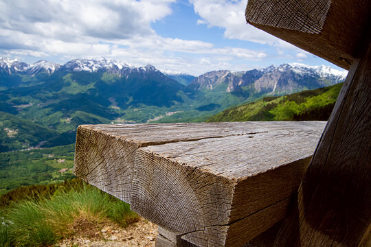 Close up of an empty weathered wooden picnic bench on top of Monte Novegno mountain. Scenic alpine landscape with blurry snow capped peaks under blue cloudy sky in Schio, Italy