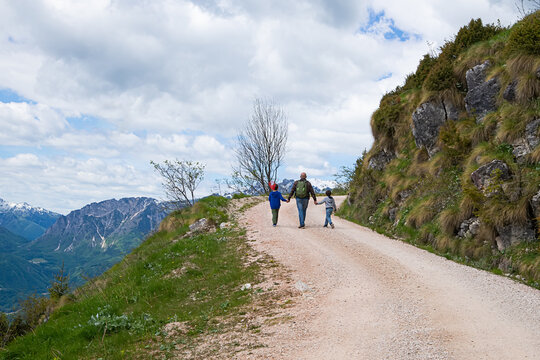 Rear view of a Caucasian father with his two young children holding hands and walking along a dirt road on Monte Novegno mountain. Active family summer vacation in Schio, Veneto, Italy