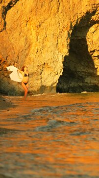 Bikini woman stands by golden cliffside cove holding a light shawl while ocean waves crash against the shore. Static wide shot.