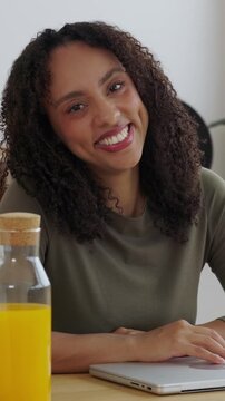 Vertical HD video of smiling young woman look at camera while working from home. Female professional using laptop in the kitchen while starting her day with orange juice