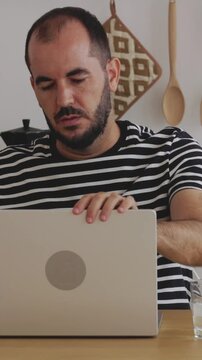 Vertical HD video of mid adult man sitting at a wooden table in a bright modern kitchen, closing his laptop after finishing work for the day and smiling confidently at the camera